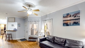 A living room with a black leather sofa, wooden flooring, a ceiling fan, and a painting of palm trees. - Sunny condo with pool, hot tub, tennis court, & gym - balcony & washer/dryer (Kissimmee)