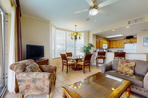 Ein Wohnzimmer mit Sofa, Couchtisch und Fernseher. Ein Esstisch mit Stühlen und einer Kristallleuchtreiter sind ebenfalls vorhanden. Das Zimmer verfügt über große Fenster und einen Deckenventilator.