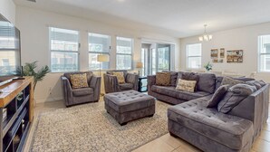 Un salon spacieux avec des canapés gris, un grand pouf et un tapis à motifs.