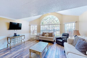 A spacious living room with a high ceiling, a large window, a flat-screen TV, a wooden coffee table, and a beige sofa. - 3BR Laguna Bay Villas, Kissimmee, near Disney (Kissimmee)