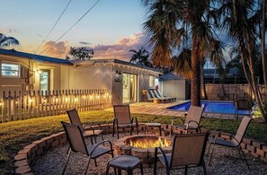A backyard with a fire pit, outdoor seating, a pool, and a house with string lights.