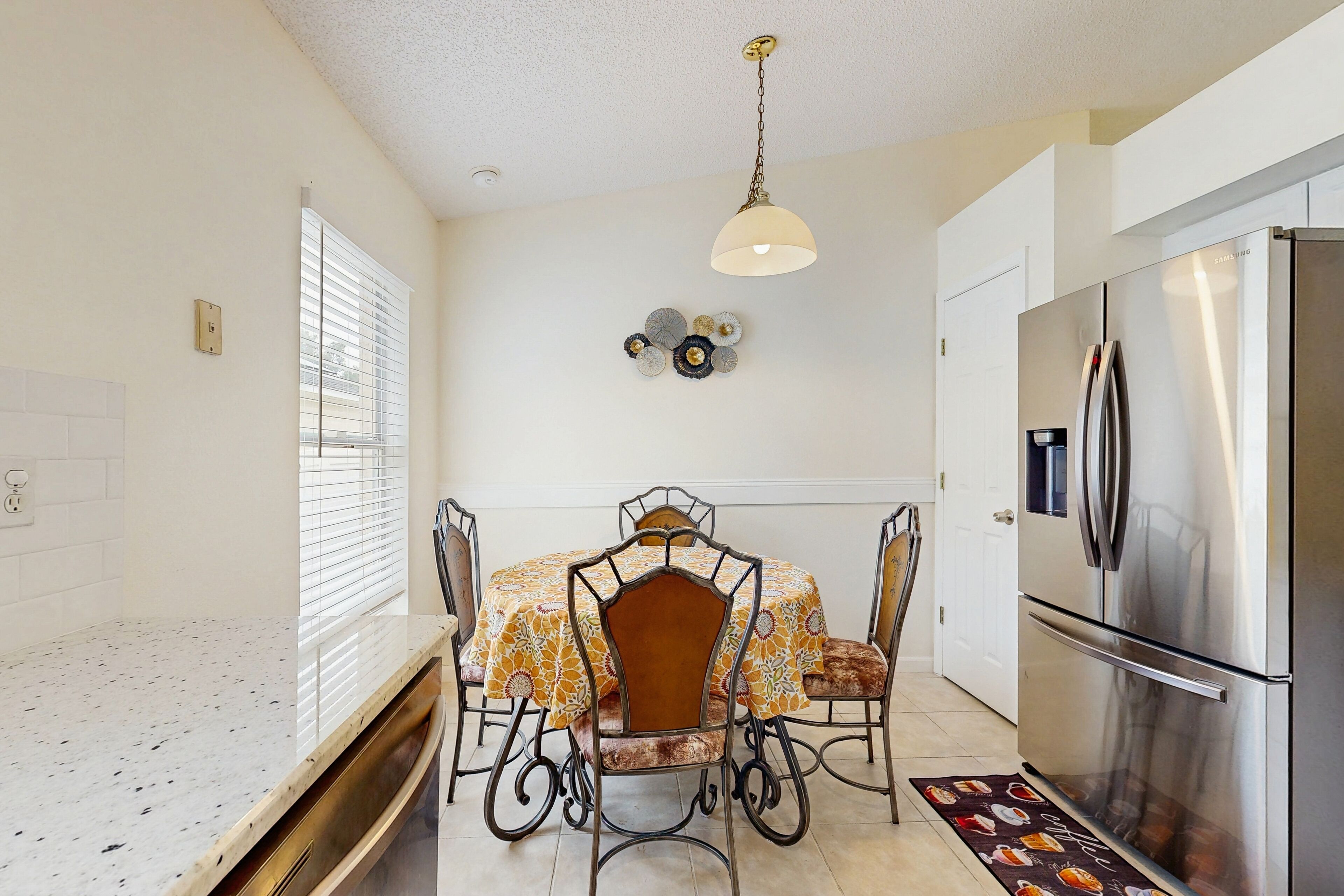 A compact kitchen with a stainless steel refrigerator, a dining table with four chairs, and a pendant light.