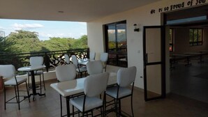 Un espacio de comedor al aire libre con mesas y sillas blancas, vistas a zonas verdes y un letrero que dice 'Estamos muy orgullosos de ti'.