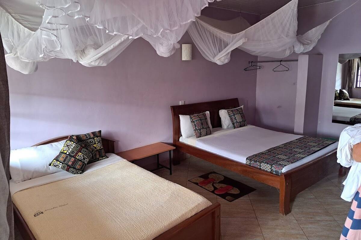 A room with two beds, one with a canopy and one without, a wooden nightstand, a small table, and a mirror on the wall.