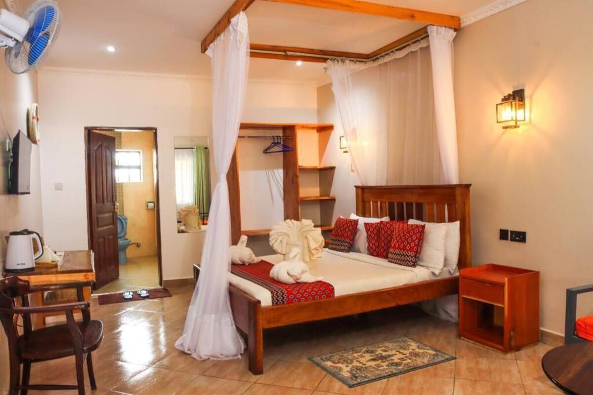 A bedroom with a wooden bed, white bedding, a bedside table, a nightstand, a chair, a fan, and a wall-mounted light fixture.