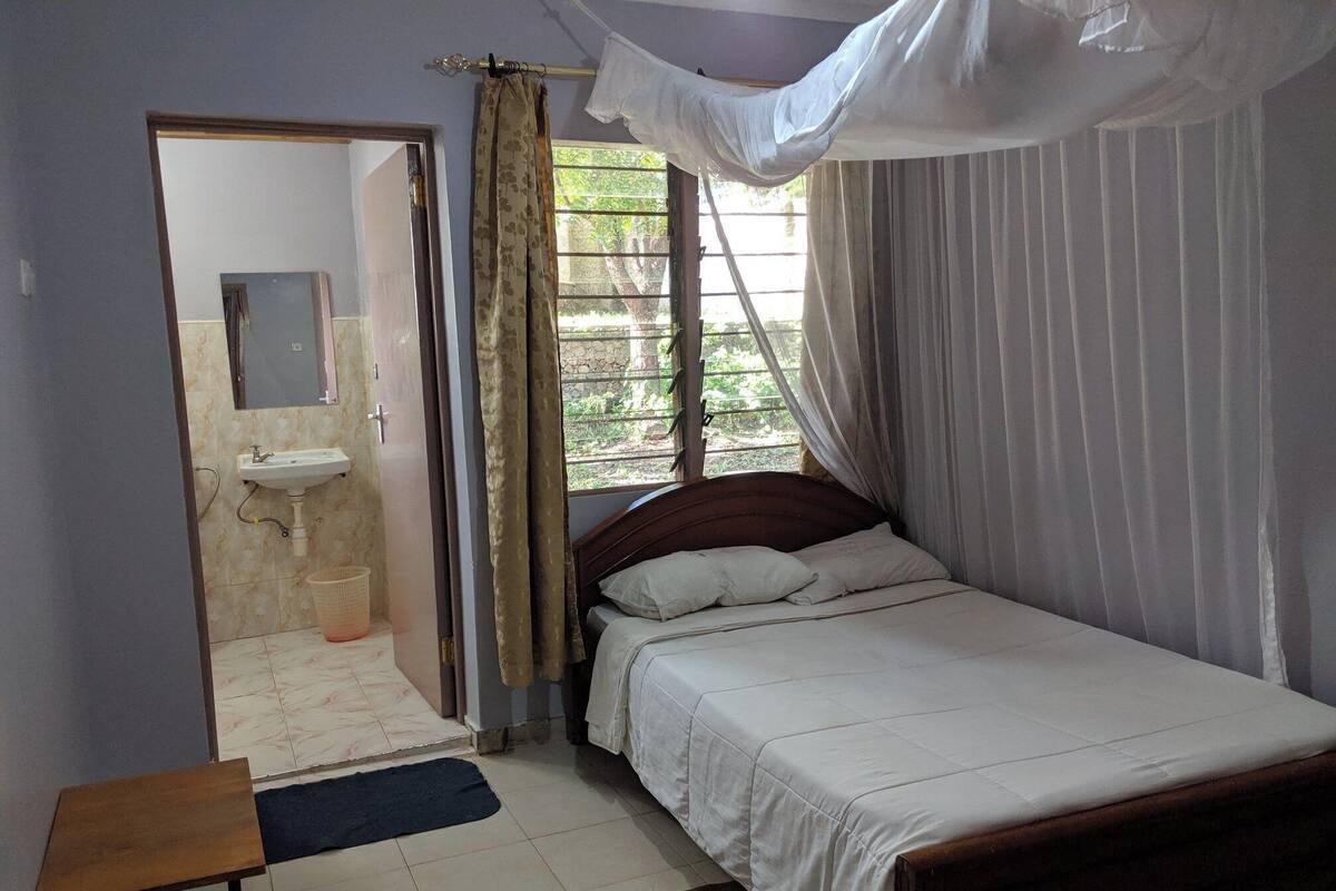 A bedroom with a bed, a bedside table, a window with curtains, a sink, and a mirror.