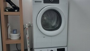 Lavatrice e asciugatrice affiancate in una lavanderia, con uno scaffale in legno sopra dove sono riposti un asciugacapelli e un flacone spray.