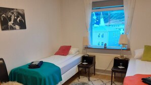 Una camera da letto piccola e ordinata con un letto, una sedia, un comodino, una lampada e una finestra vista edifici.