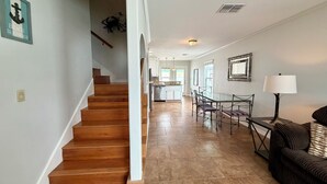 A spacious living area with a wooden staircase, a dining table with chairs, a sofa, and a kitchen in the background. - GS6 Shared Heated Pool, Walk to the Beach, Golf Cart Access (Port Aransas)