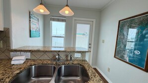 A kitchen with granite countertops, stainless steel sinks, pendant lighting, a window with blinds, and a framed map on the wall. - GS6 Shared Heated Pool, Walk to the Beach, Golf Cart Access (Port Aransas)
