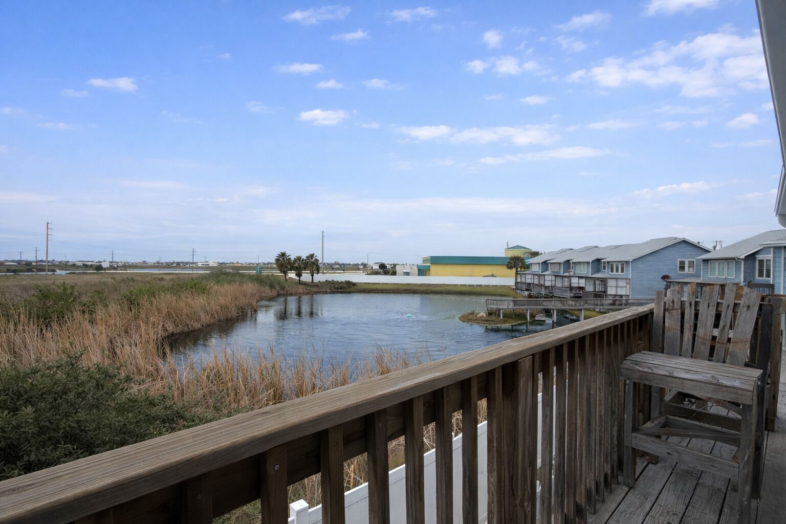 A wooden deck overlooking a calm body of water with a marshy area in the foreground and a row of blue houses in the background.