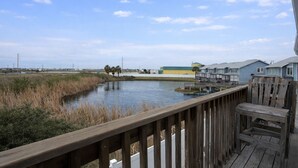 A wooden deck overlooking a calm body of water with a marshy area in the foreground and a row of blue houses in the background. - GS6 Shared Heated Pool, Walk to the Beach, Golf Cart Access (Port Aransas)