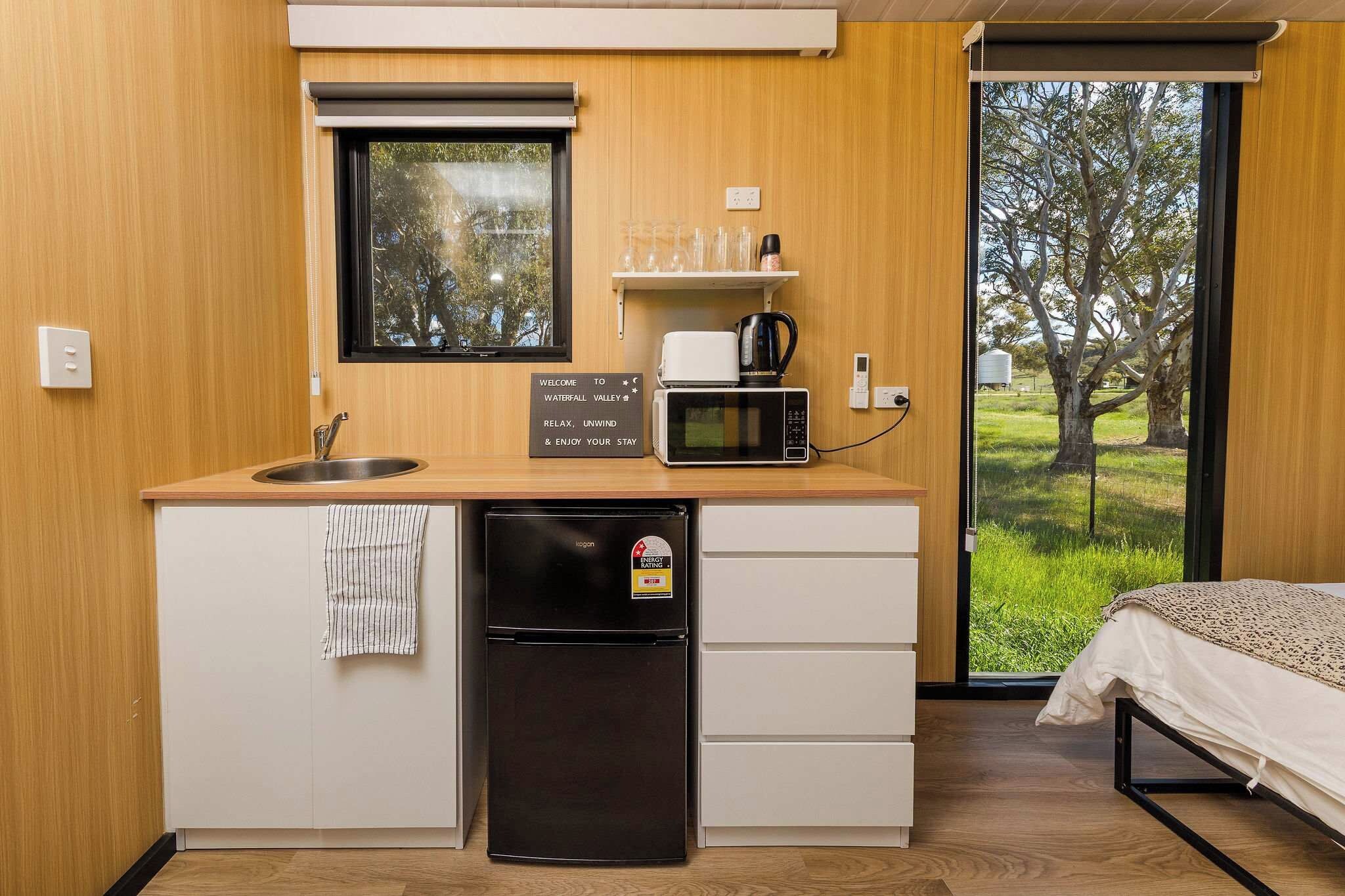 A compact kitchenette with a sink, a black refrigerator, a microwave, and a coffee maker. There is a sign above the microwave that reads 'Welcome to Waterfall Valley.'