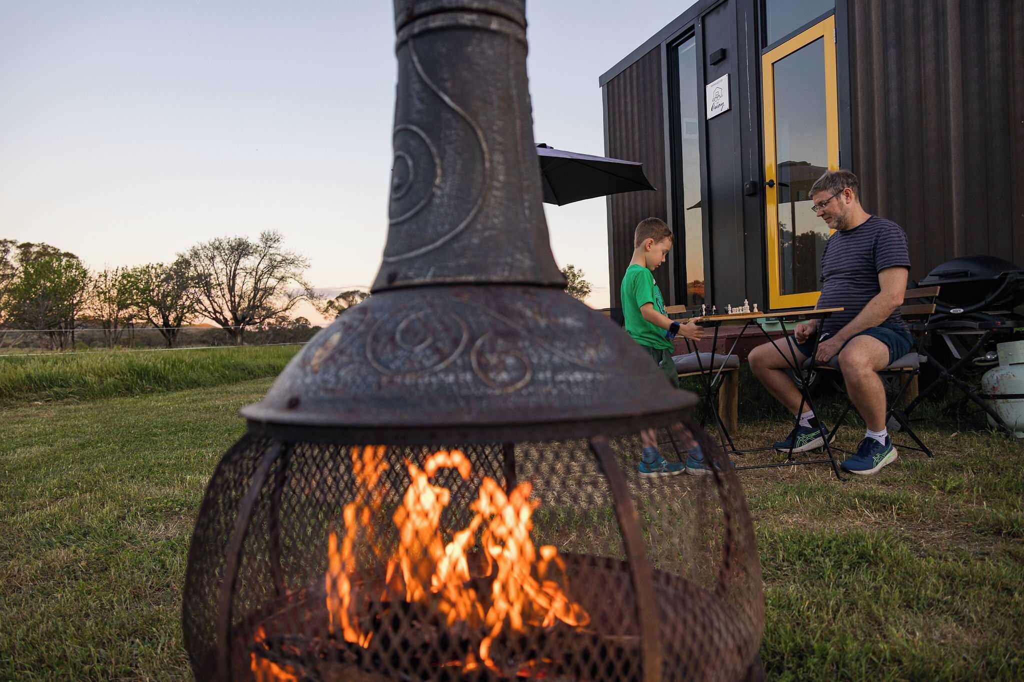 A metal fire pit with flames, a wooden table with chess pieces, and two people seated at the table.