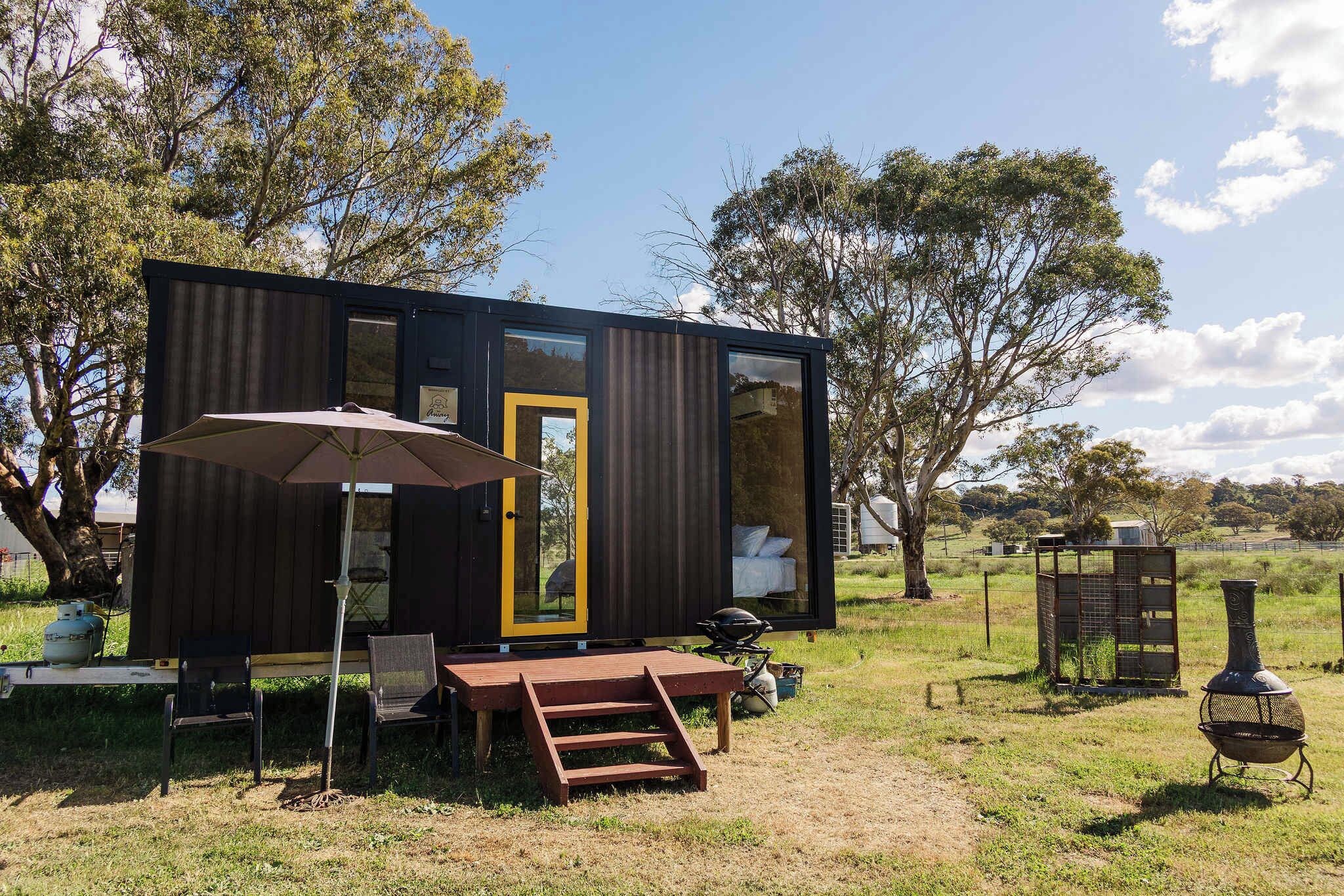 Una casa móvil moderna de color oscuro con una puerta amarilla, un patio con mesa y sillas, y una parrilla de barbacoa en un área con césped.