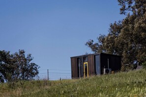 A small, dark-colored building with a yellow door, surrounded by grass and trees, with a clear sky above. - Waterfall Valley by Tiny Away (Gowang)