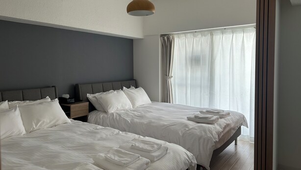 A hotel room with two beds, a wooden headboard, a bedside table, a nightstand, and a large window with sheer curtains. - TOWN ROYAL 601 (Sapporo)