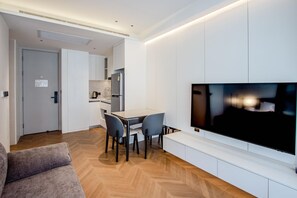 Ein modernes Wohnzimmer mit großem Flachbildfernseher an der Wand, einem Essbereich mit Tisch und Stühlen sowie einer Küchenzeile inklusive Kühlschrank und Wasserkocher.