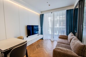 A modern living room with a flat-screen TV mounted on the wall, a sofa, a dining table with chairs, and a large window with sheer curtains.