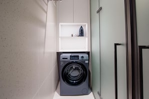 A compact laundry room with a front-loading washing machine, a small shelf above it, and a door to the right.