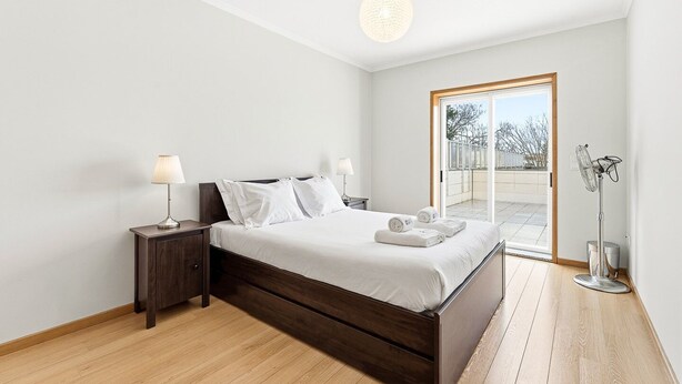 A modern bedroom with a wooden bed, two bedside tables with lamps, a large window with a view of the outside, and a ceiling light. - GuestReady - Charming Porto retreat with terrace (Boavista)