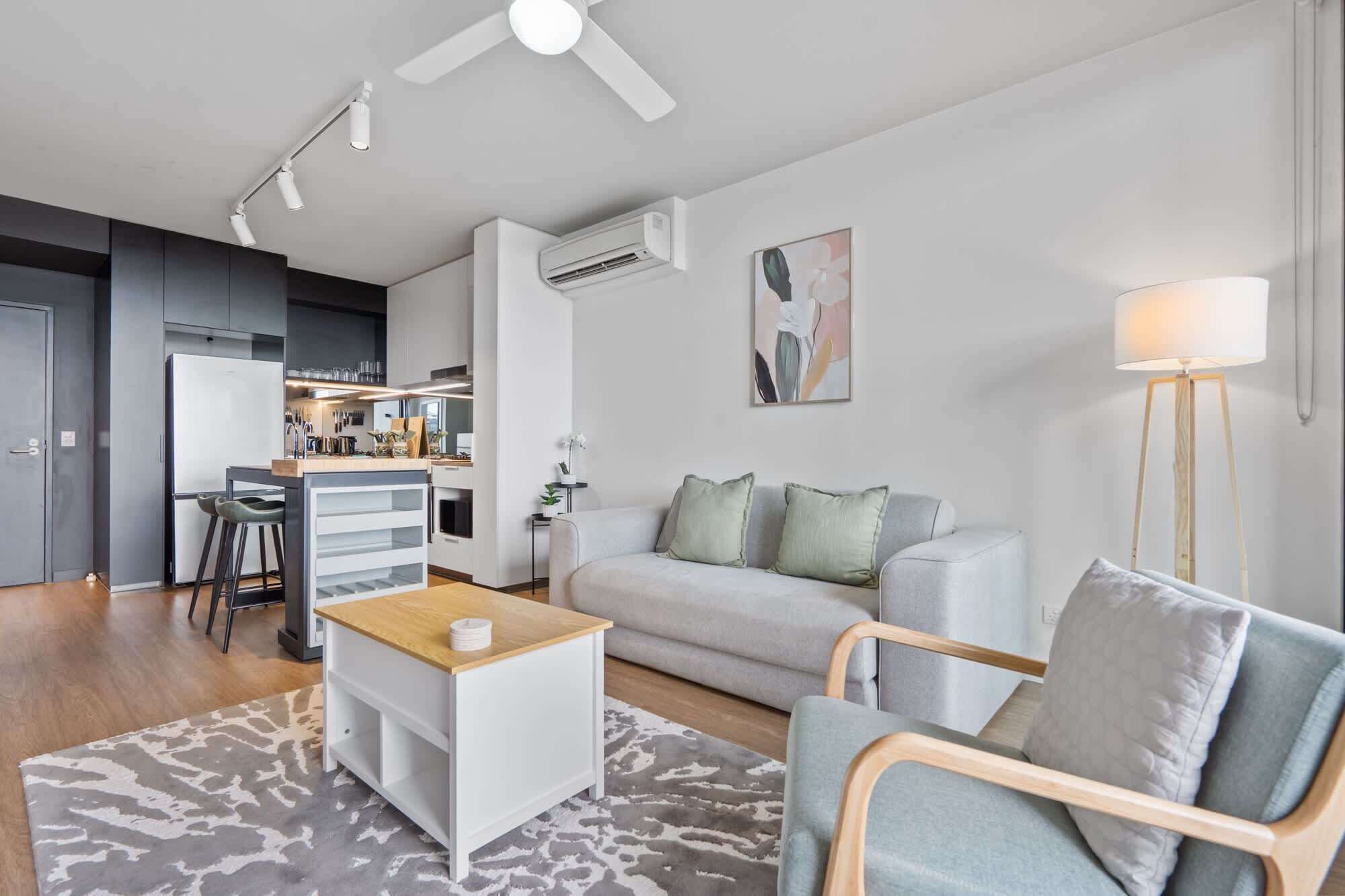 A modern living room with a grey sofa, a wooden coffee table, and a white side table. There is a kitchen area with a fridge, stove, and bar stools. A ceiling fan and an air conditioning unit are visible.