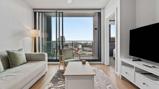 A modern living room with a sofa, a coffee table, a TV, and a balcony with a city view. - Bright 2-Bed Apartment in Bowen Hills (Bowen Hills)