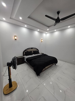 A bedroom with a bed, a standing fan, recessed ceiling lights, and a ceiling fan.