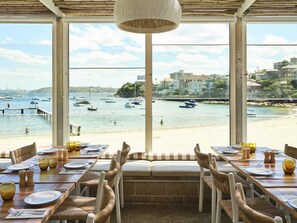 A dining area with a view of a beach and boats. Tables are set with plates, glasses, and cutlery. - Sunny Manly Apartment Steps from Beach & Cafes (Manly)