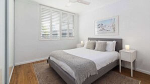 Ein Schlafzimmer mit einem Bett, zwei Nachttischen, einem Deckenventilator, einem Fenster mit Jalousien, einem gerahmten Bild an der Wand und einem Teppich auf dem Boden.