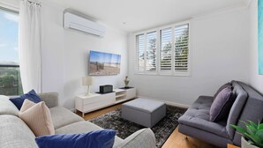 Ein modernes Wohnzimmer mit einem grauen Sofa, einem grauen Sessel und einem weißen Beistelltisch mit Lampe. Ein großes Fenster mit Jalousien, ein Flachbildfernseher und eine kleine Pflanze auf einem Holztisch sind ebenfalls vorhanden.