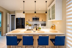 Une cuisine moderne dotée d’un îlot central, de tabourets de bar, d’appareils électroménagers en acier inoxydable et de luminaires suspendus.