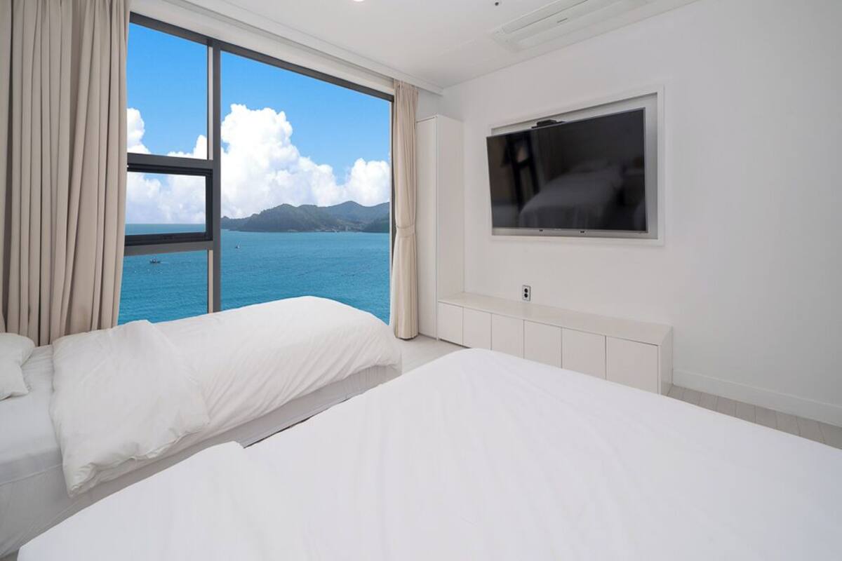 A hotel room with a large window offering a sea view, a bed, a TV, and a white cabinet.