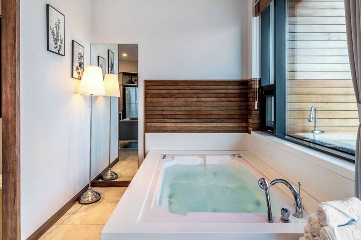 A modern bathroom with a large bathtub, a wooden headboard, a window with blinds, and a floor lamp.