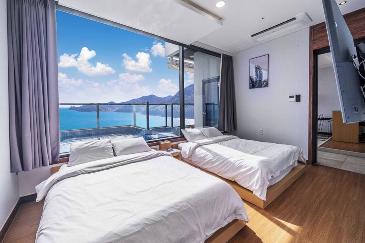A modern hotel room with two beds, large windows offering a sea view, and a balcony with a glass barrier.