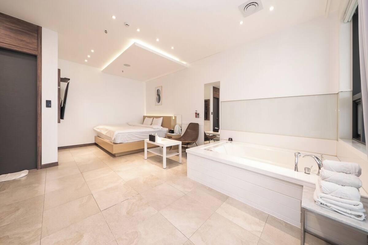 A modern bedroom with a large bed, a sitting area, and a separate bathroom with a bathtub and a large mirror.