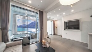 Ett modernt hotellrum med ett stort fönster som erbjuder en vacker utsikt över berg och vatten, en bekväm soffa, ett litet bord med diverse bekvämligheter och en platt-TV som är monterad på väggen.