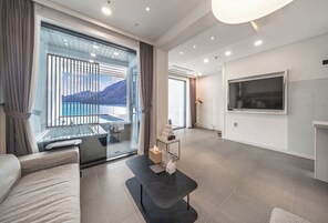 Habitación de hotel moderna con un gran ventanal que ofrece vistas panorámicas de montañas y agua, un sofá cómodo, una mesita con artículos de cortesía y un televisor de pantalla plana montado en la pared.