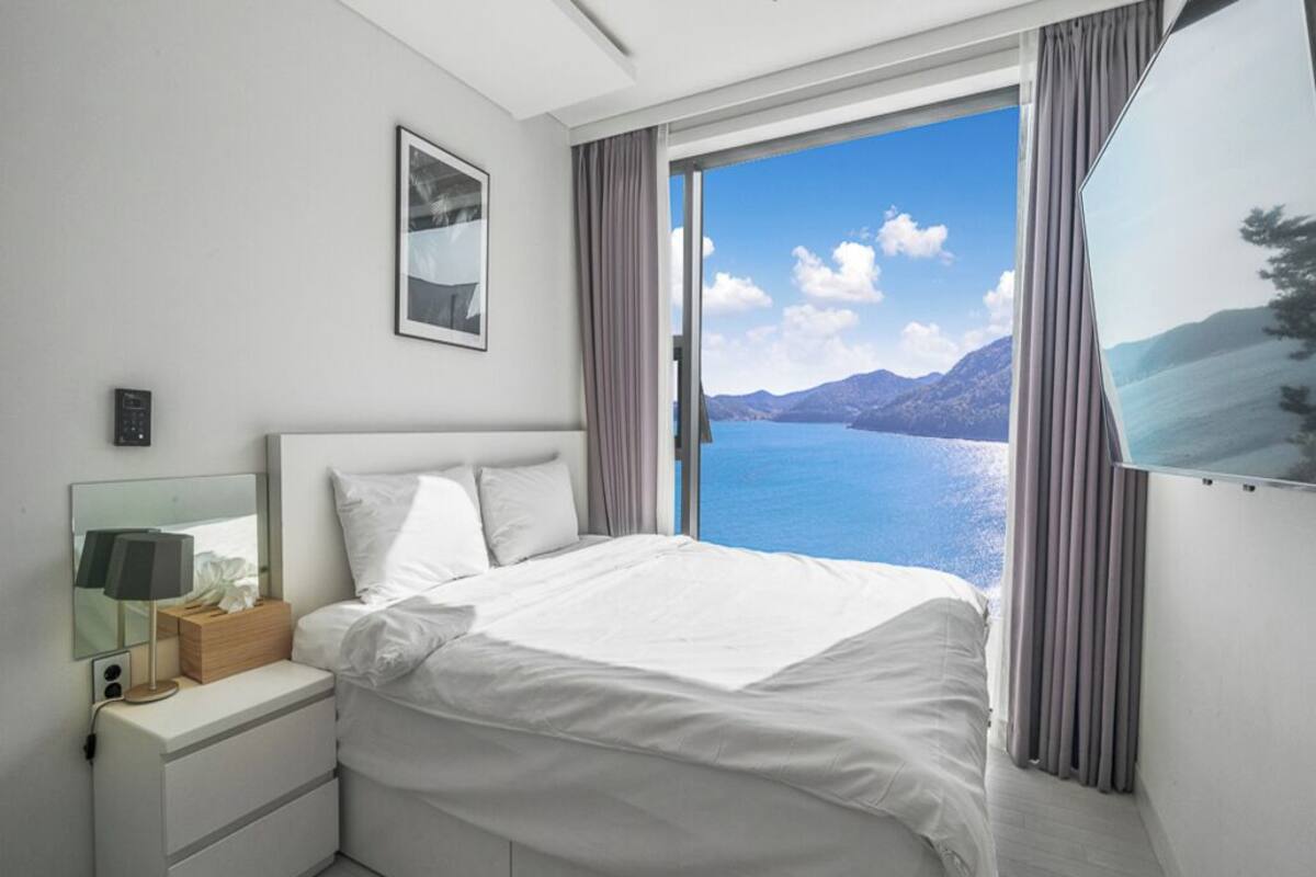 A modern hotel room with a large bed, a bedside table with a lamp, a mirror, and a view of the sea and mountains.