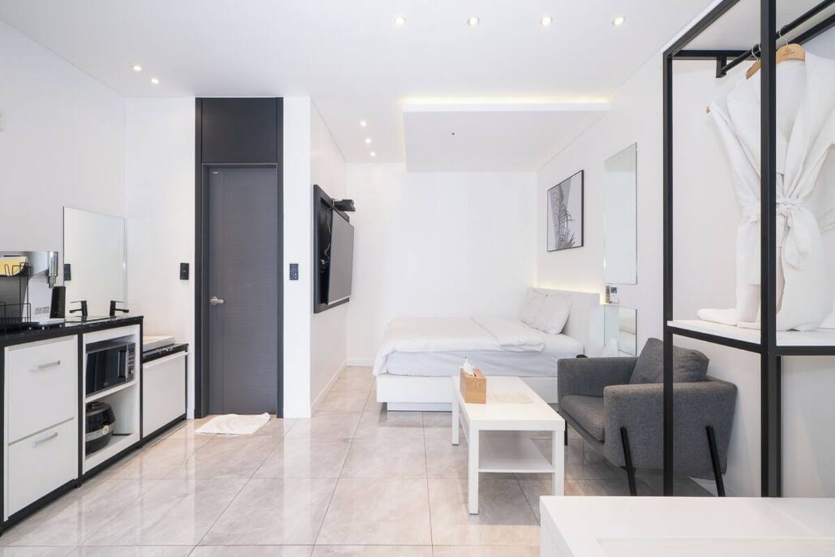 A modern, minimalist hotel room with a kitchenette, a bed, a gray sofa, and a white coffee table.