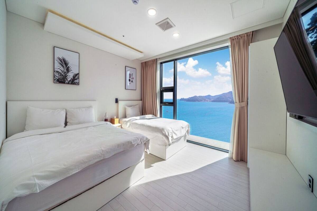 A hotel room with two beds, a large window with a view of the sea, and a flat-screen TV.