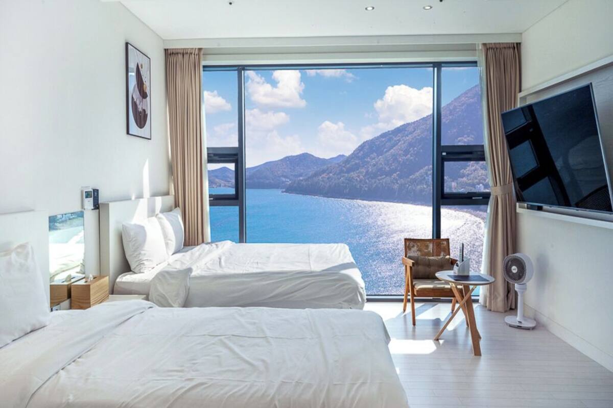 A hotel room with two beds, a large window with a mountain and lake view, a flat-screen TV, and a small table with a fan.