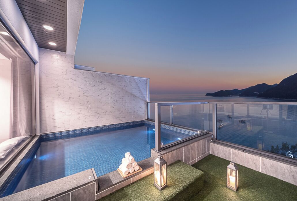 Piscina infinita con vistas despejadas al mar y a las montañas, barandilla de cristal, y una pequeña zona con césped artificial y faroles.