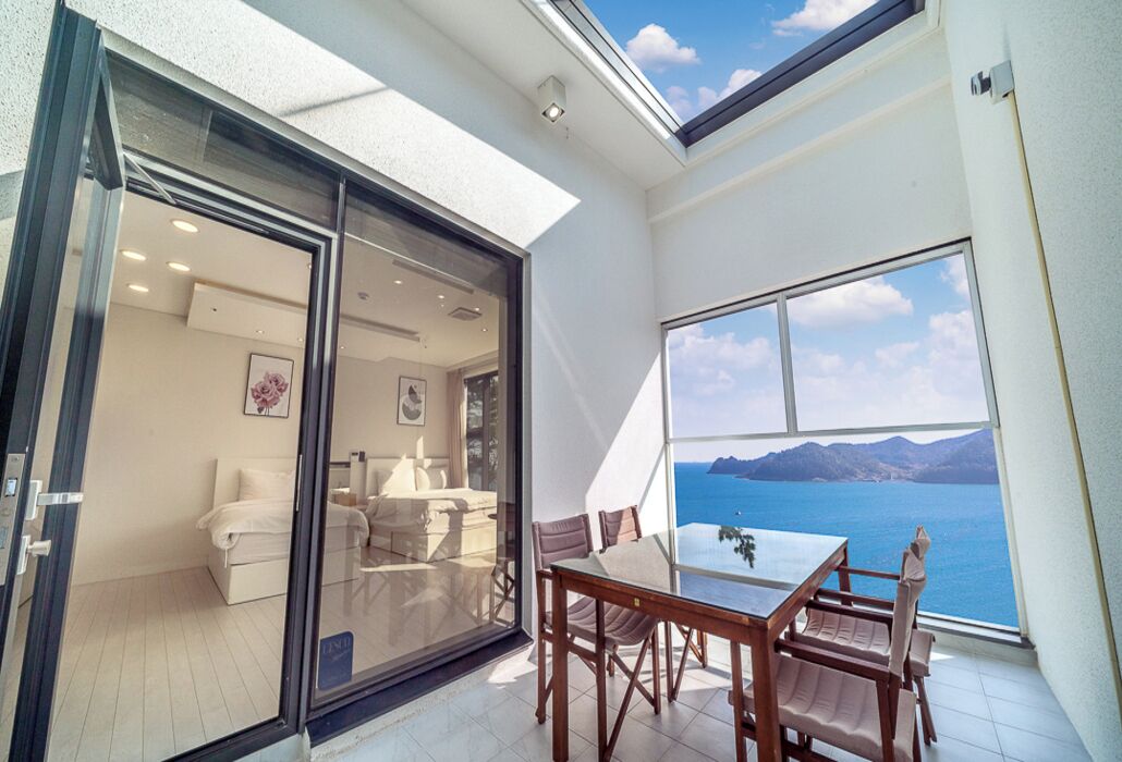 Un balcón con mesa y sillas, un ventanal corredero que da a una habitación con cama y sofá, y un gran ventanal con vistas al mar y las montañas.
