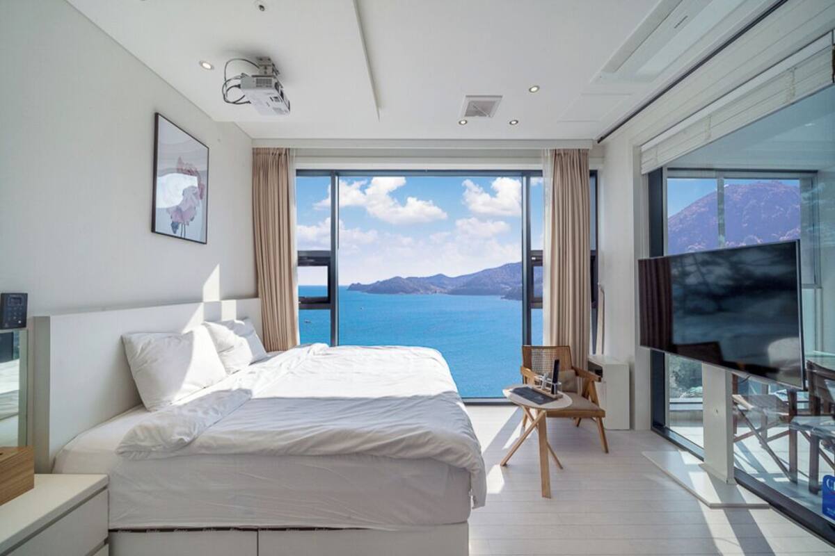 A modern bedroom with a large bed, a TV, and a view of the sea.