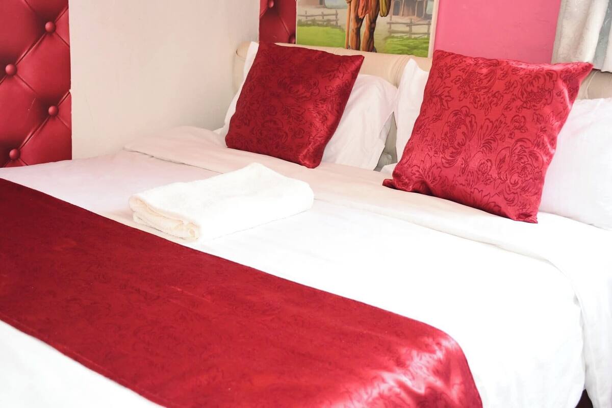 Una cama bien hecha con sábanas blancas y almohadas rojas, una manta roja y un cabecero capitonado.
