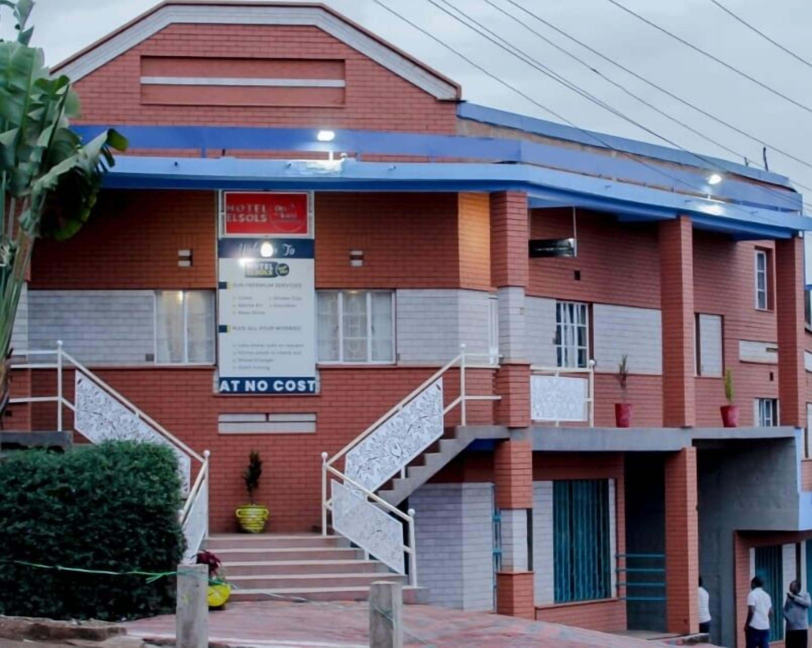 A two-story building with a red brick facade, a blue roof, and a staircase leading to the entrance. There is a sign that reads "AT NO COST" and another that says "HOTEL SERVICES."