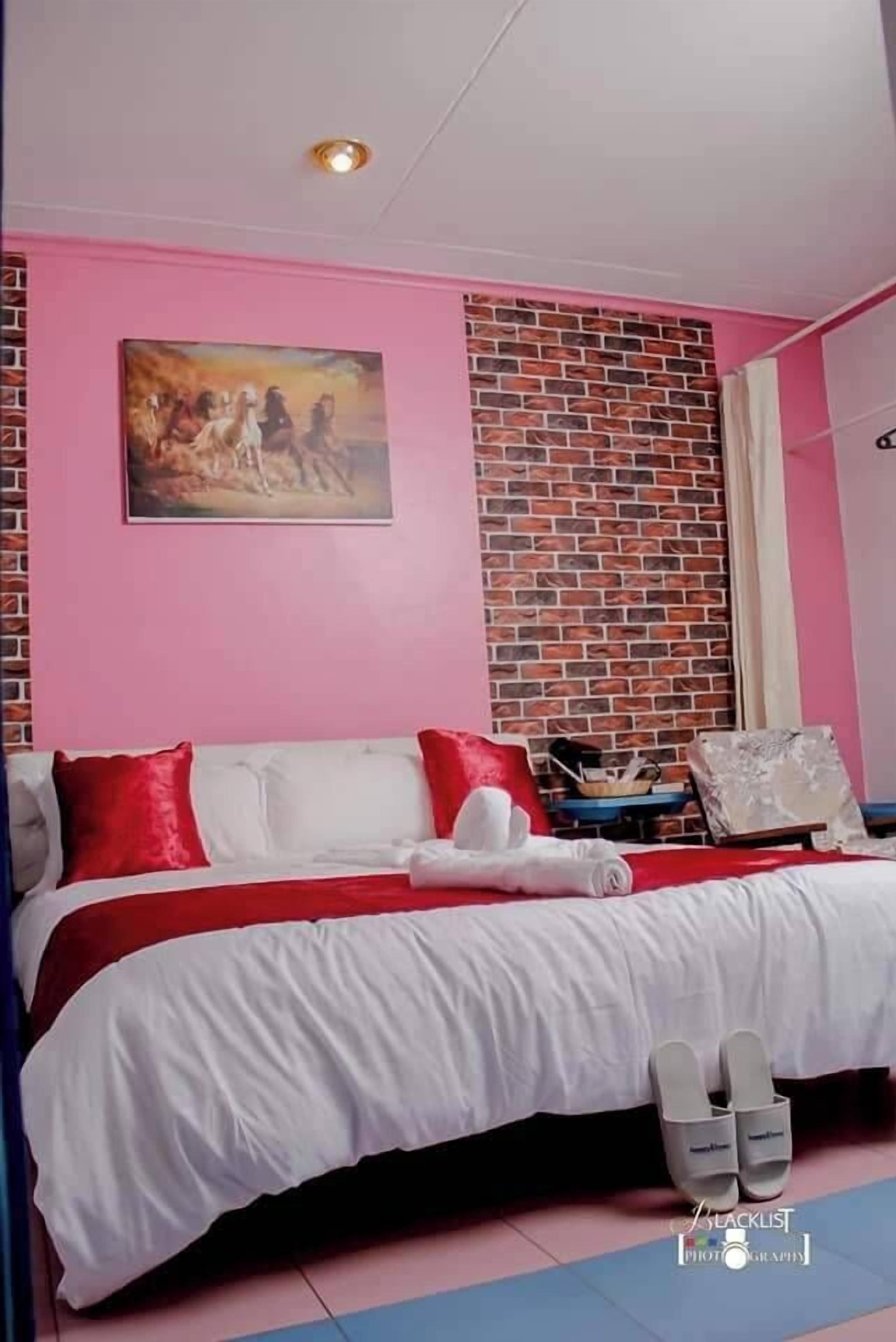 A bedroom with a brick-patterned accent wall, a painting above the bed, and a pair of slippers on the floor.