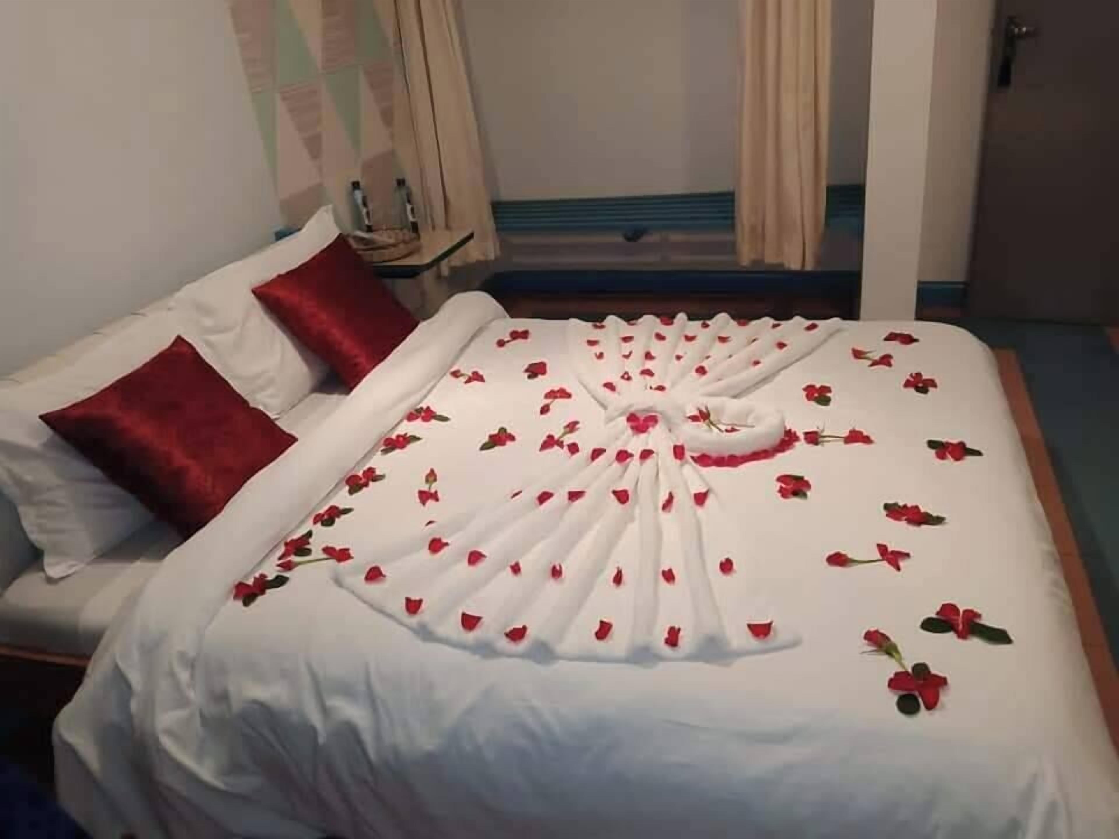 Een net opgemaakt bed met een witte spreideken en rode kussens, een klein tafeltje met een fles en een glas, en een deur op de achtergrond.