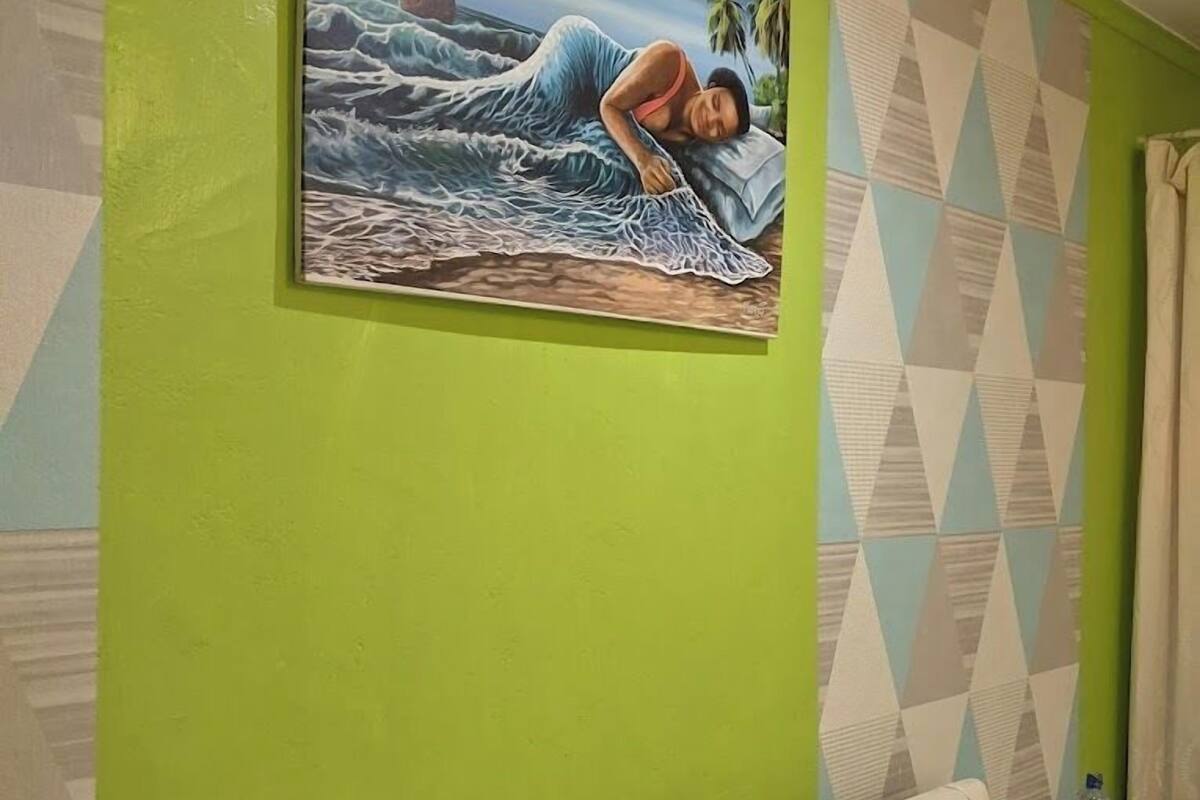 Una habitación con pared verde, un cuadro de una mujer en un bote, un papel tapiz estampado y un sofá blanco con almohadas rojas.